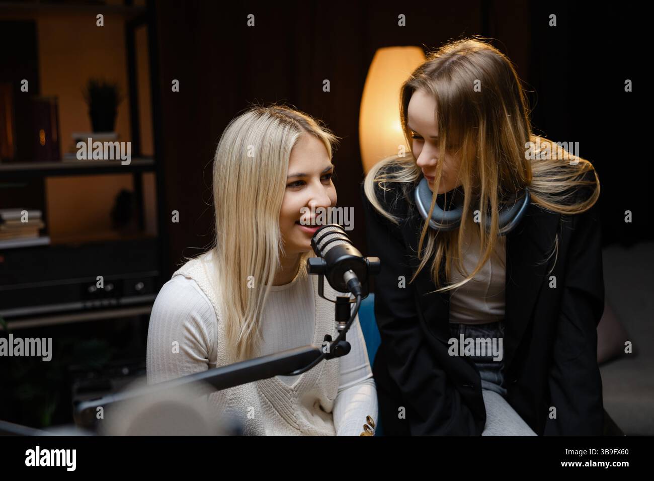 Zwei weibliche Podcaster, die Audioinhalte erstellen und gleichzeitig professionelle Mikrofone in einem gemütlichen Heimstudio verwenden Stockfoto
