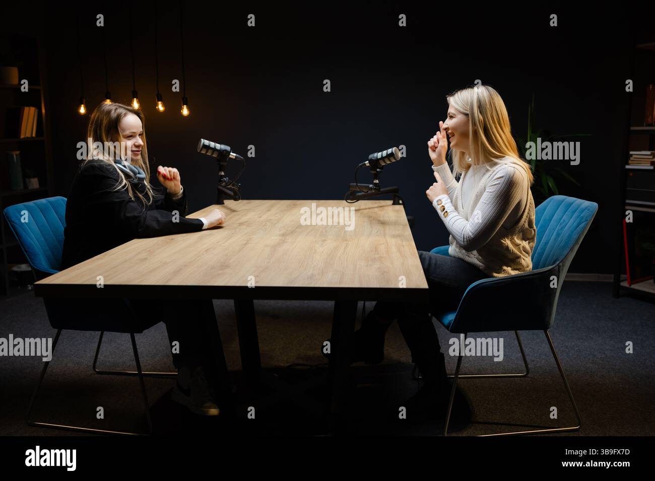 Zwei Frauen nehmen in einem professionellen Studio einen Audio-Podcast auf, sitzen mit Mikrofonen an einem Tisch, diskutieren und Gesten geben Stockfoto