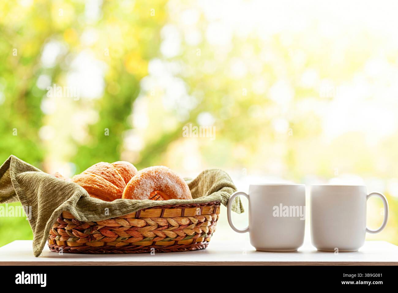 Ein Korb voller frischer Croissants und süßer Donuts steht neben zwei weißen Kaffeetassen auf einem Tisch und bietet einen herrlichen Blick auf den üppig grünen Garten Stockfoto