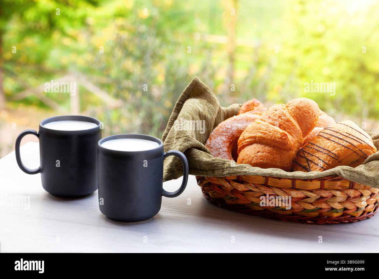 Zwei dampfende Tassen Milch und ein Korb mit frisch gebackenen Croissants und Gebäck sitzen auf einem Tisch im Freien, was eine friedliche und einladende Frühstücksszene schafft Stockfoto