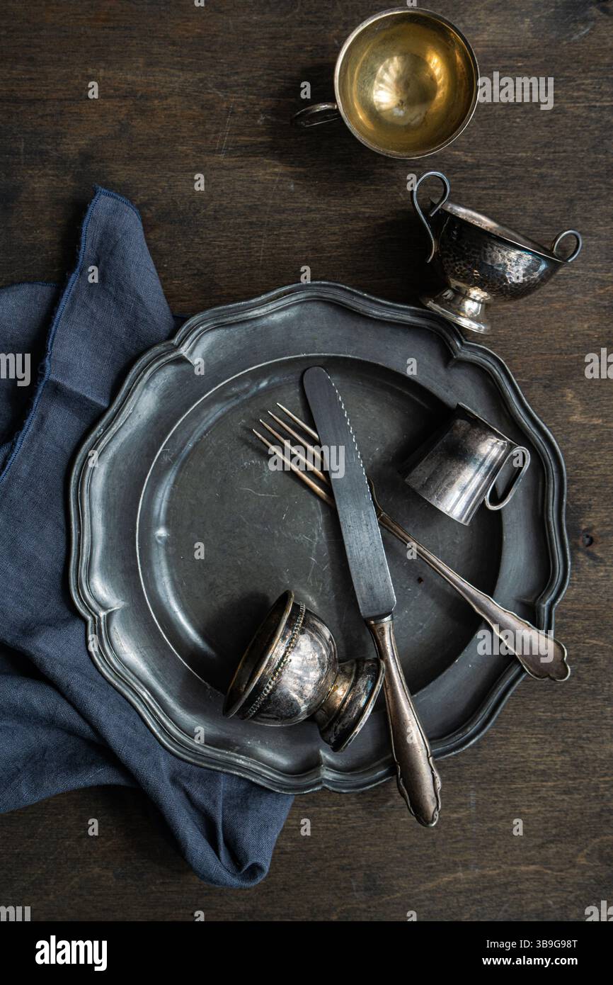 Ein rustikales Arrangement von Vintage-Metallgeschirr mit Teller, Gabel, Messer und zwei kleinen Tassen auf dunkler Holzoberfläche, ergänzt durch ein blaues n Stockfoto