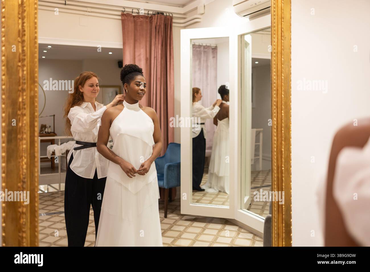Eine Braut steht vor einem Spiegel in einem Umkleideraum, während eine Stylistin ihr weißes Hochzeitskleid anpasst und die Spannung und Eleganz des M widerspiegelt Stockfoto