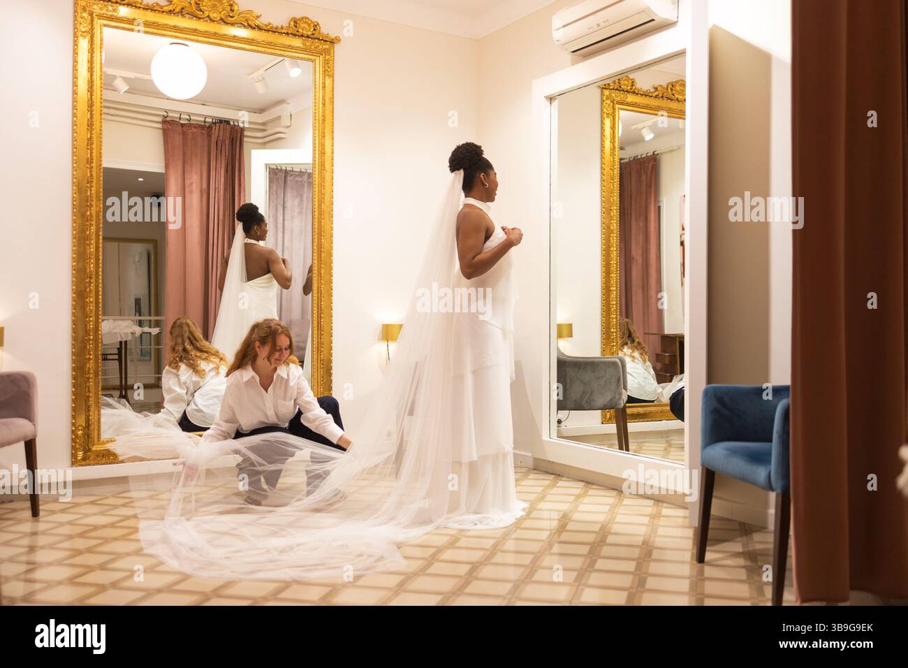 Braut steht elegant in einer Garderobe, ihr langer Schleier wird von einer Assistentin angepasst. Das Zimmer verfügt über große Spiegel und einen goldgerahmten Wandspiegel, Stockfoto