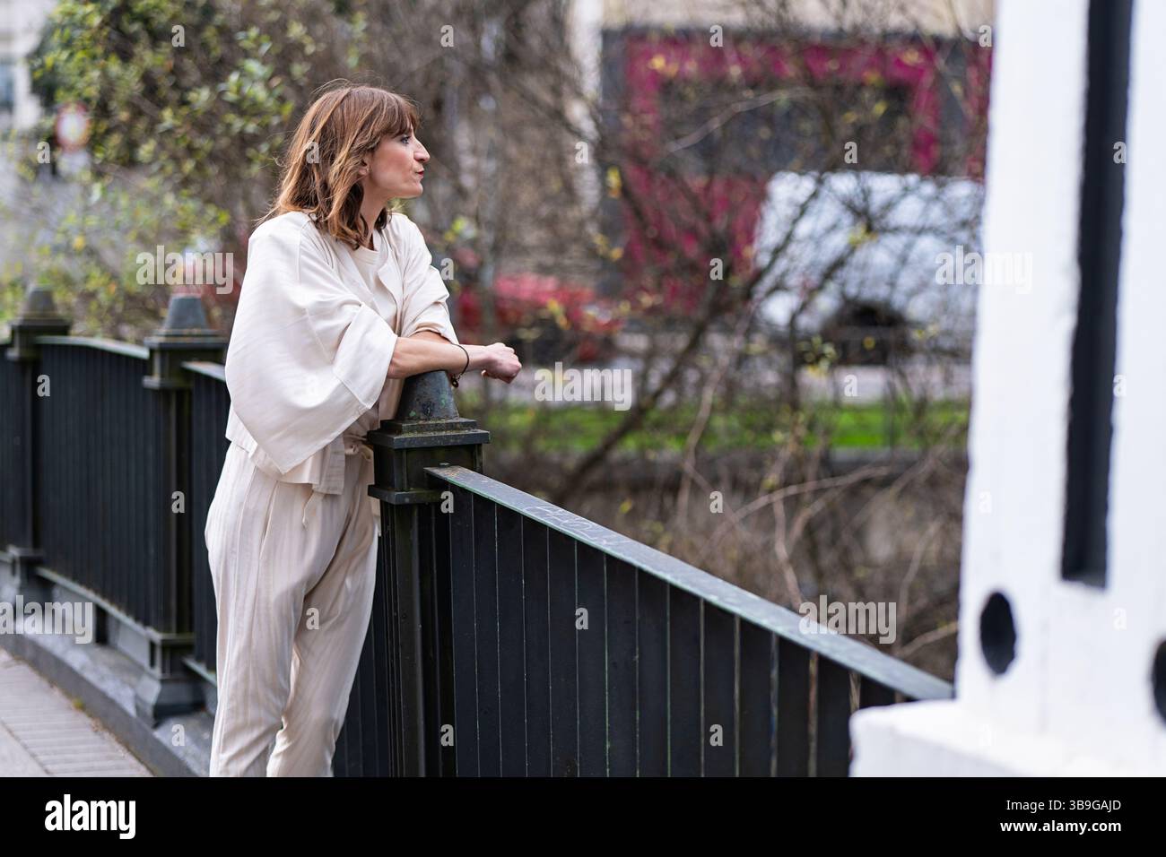 Eine modische Frau in einem schicken Outfit lehnt sich nachdenklich auf ein Brücken-Geländer der Stadt. Der urbane Hintergrund und ihre entspannte Pose wecken ein Gefühl der Besinnung Stockfoto