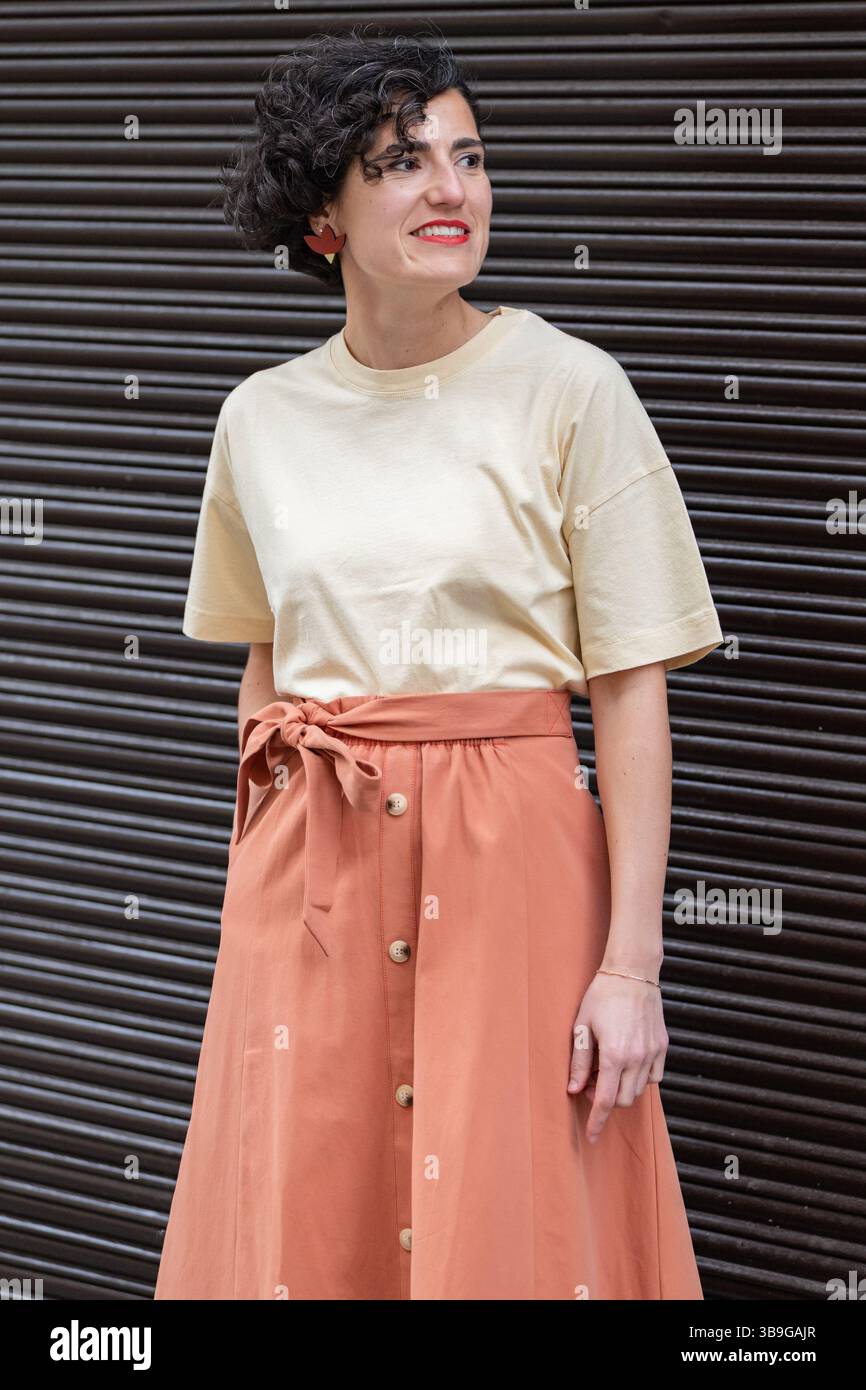 Eine modische Frau in einem beigefarbenen T-Shirt und einem orangefarbenen Rock mit Schleife steht vor einem dunklen Hintergrund. Ihr kurzes lockiges Haar und die roten Ohrringe ergänzen sich Stockfoto