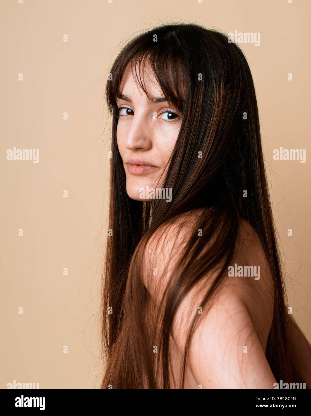 Ein Porträt einer jungen Frau mit langen braunen Haaren, die in einem Atelier auf beigefarbenem Hintergrund die Kamera über ihre Schulter schaut. Ihr Blick ist subtil und fügt de hinzu Stockfoto