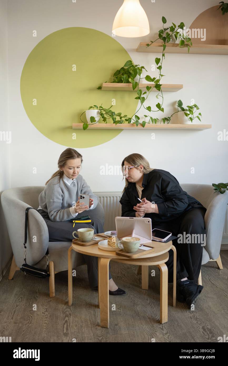 Zwei Frauen unterhalten sich in einem gemütlichen Café. Umgeben von Kaffee, Gebäck und Grün, genießen sie eine entspannte und ungezwungene Atmosphäre Stockfoto