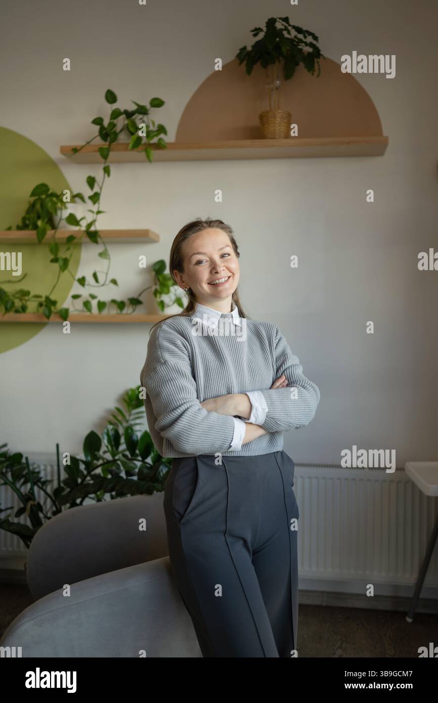 Eine lächelnde Frau steht selbstbewusst in einem gemütlichen Café, umgeben von üppig grünen Pflanzen. Sie trägt einen grauen Pullover und ein weißes Hemd, was eine entspannte und doch entspannte Farbe verkörpert Stockfoto
