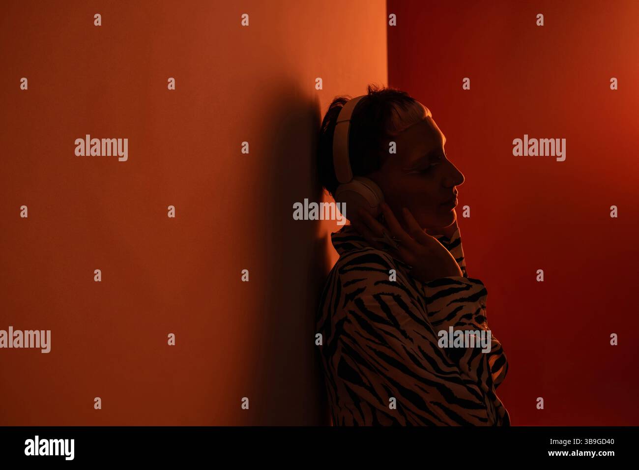 Junge trendige Frau in einem Zebramuster-Outfit, lehnt sich an eine Wand und genießt Musik über Kopfhörer, beleuchtet durch warmes, sanftes Licht, Reflektoren Stockfoto