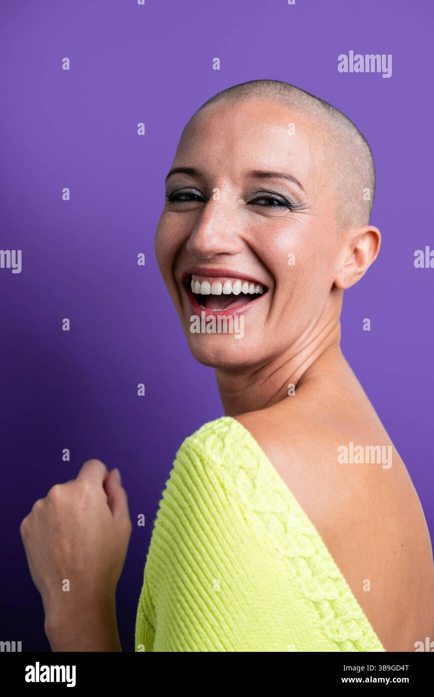 Ein lebendiges Porträt einer Frau mittleren Alters mit einem rasierten Kopf, die hell vor einem violetten Hintergrund lächelt. Das Bild fängt das Wesen der Freude ein, Betrüger Stockfoto