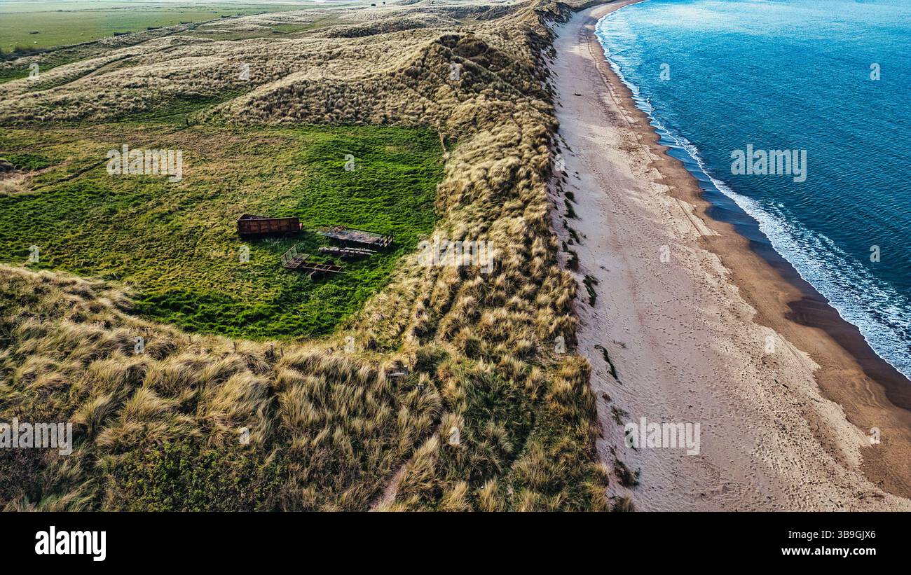 Blick aus der Vogelperspektive auf einen Sandstrand, der auf das blaue Meer trifft, umgeben von grasbewachsenen Dünen und einem grünen Feld mit rostiger Farm-Ausrüstung am Link House Beach, Großbritannien. Stockfoto