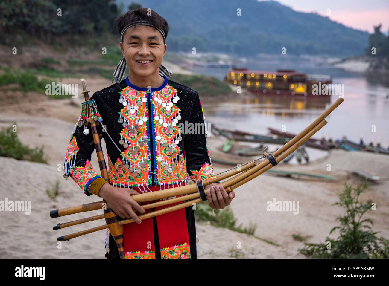 Junger Mann mit traditioneller Bambus-Mundharmonika, hinter ihm das Boutique-Kreuzfahrtschiff Anouvong (Heritage Line), das auf einer Sandbank am oberen M ankert Stockfoto