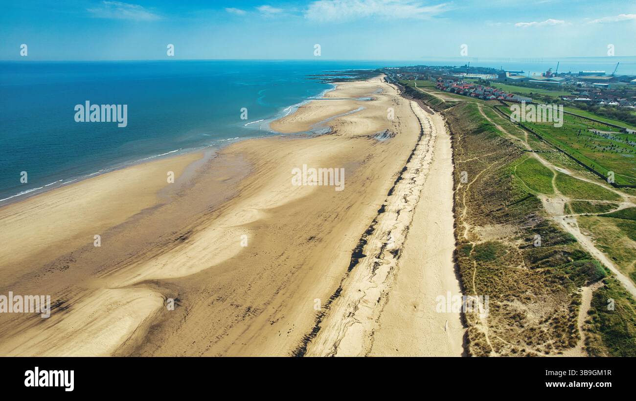 Aus der Vogelperspektive auf einen Sandstrand, der auf das blaue Meer trifft, neben grasbewachsenen Dünen und einer Stadt am Steetley Pier, Großbritannien. Stockfoto