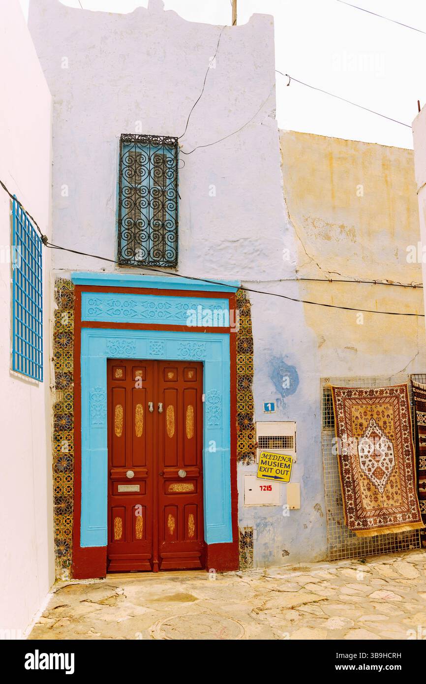 Traditionelles Haus mit farbenfroher Holztür mit arabischen Schnitzereien und gewebten Teppichen zum Verkauf an der Hauswand in Hammamet, Tunesien Stockfoto