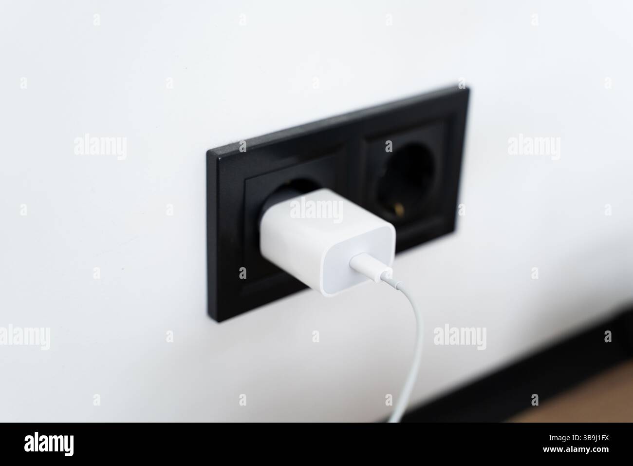 Minimalistisches Bild eines weißen USB-Ladegeräts, das in eine schwarze Doppelsteckdose an einer glatten weißen Wand gesteckt wird, veranschaulicht Technologie und täglichen Nutzen Stockfoto