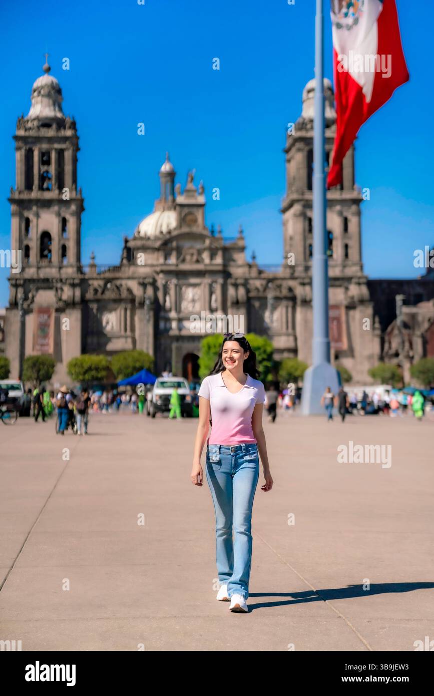Lächelnde junge Latina-Frau in lässigem rosa-weißem Hemd mit Jeans, die selbstbewusst durch den historischen Zocalo von Mexiko-Stadt unter klarem blauen Himmel spazieren. Stockfoto
