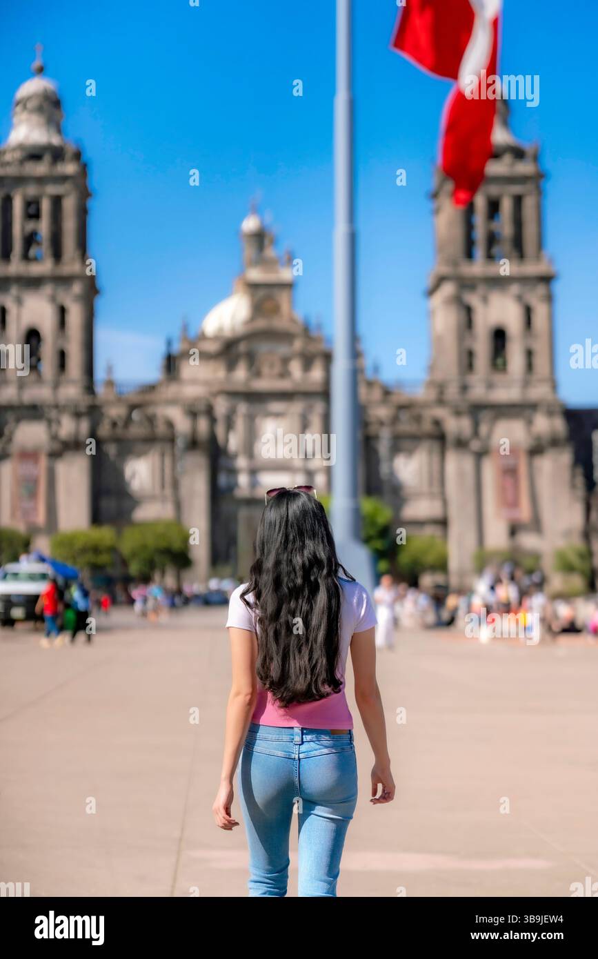 Unwiedererkennbare junge Latina-Frau in lässigem rosa-weißem Hemd mit Jeans, die selbstbewusst unter klarem Blau durch den historischen Zocalo von Mexiko City spazieren Stockfoto