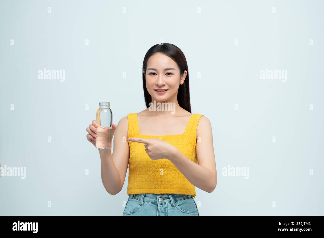 Lächelnde junge Frau in lässiger Kleidung, die auf eine klare Wasserflasche zeigt Stockfoto