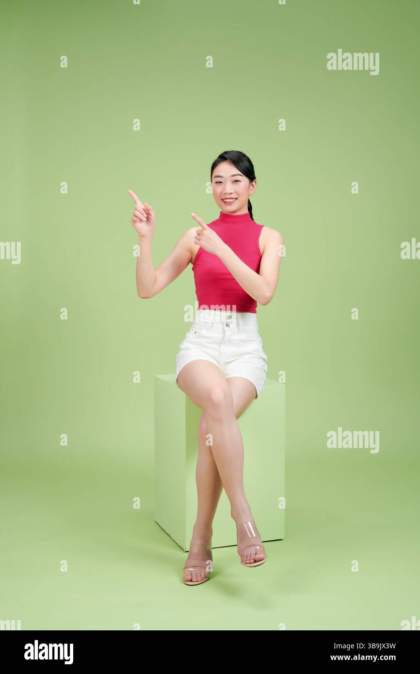 Fröhliche Frau in lässigem Outfit, die mit beiden Händen zeigt und lächelt, während sie auf grünem Hintergrund sitzt. Stockfoto