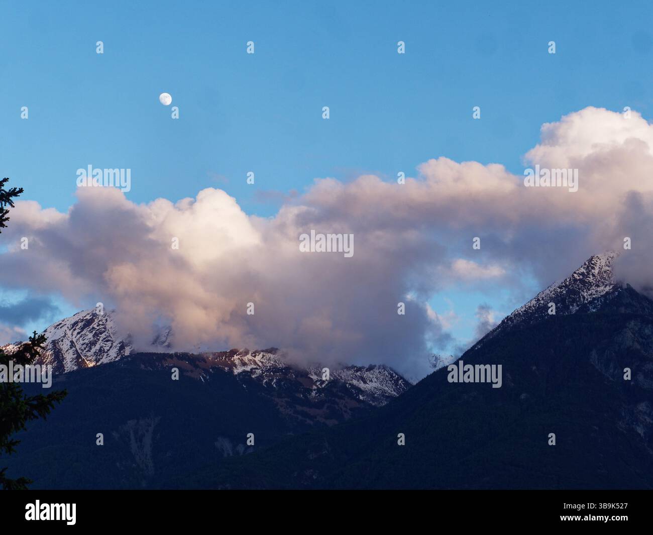 Mond hoch in einem blauen Himmel über weißen Wolken, mit schneebedeckten Bergen darunter. Aosta Valley, NW Italien. Mai 2025 Stockfoto