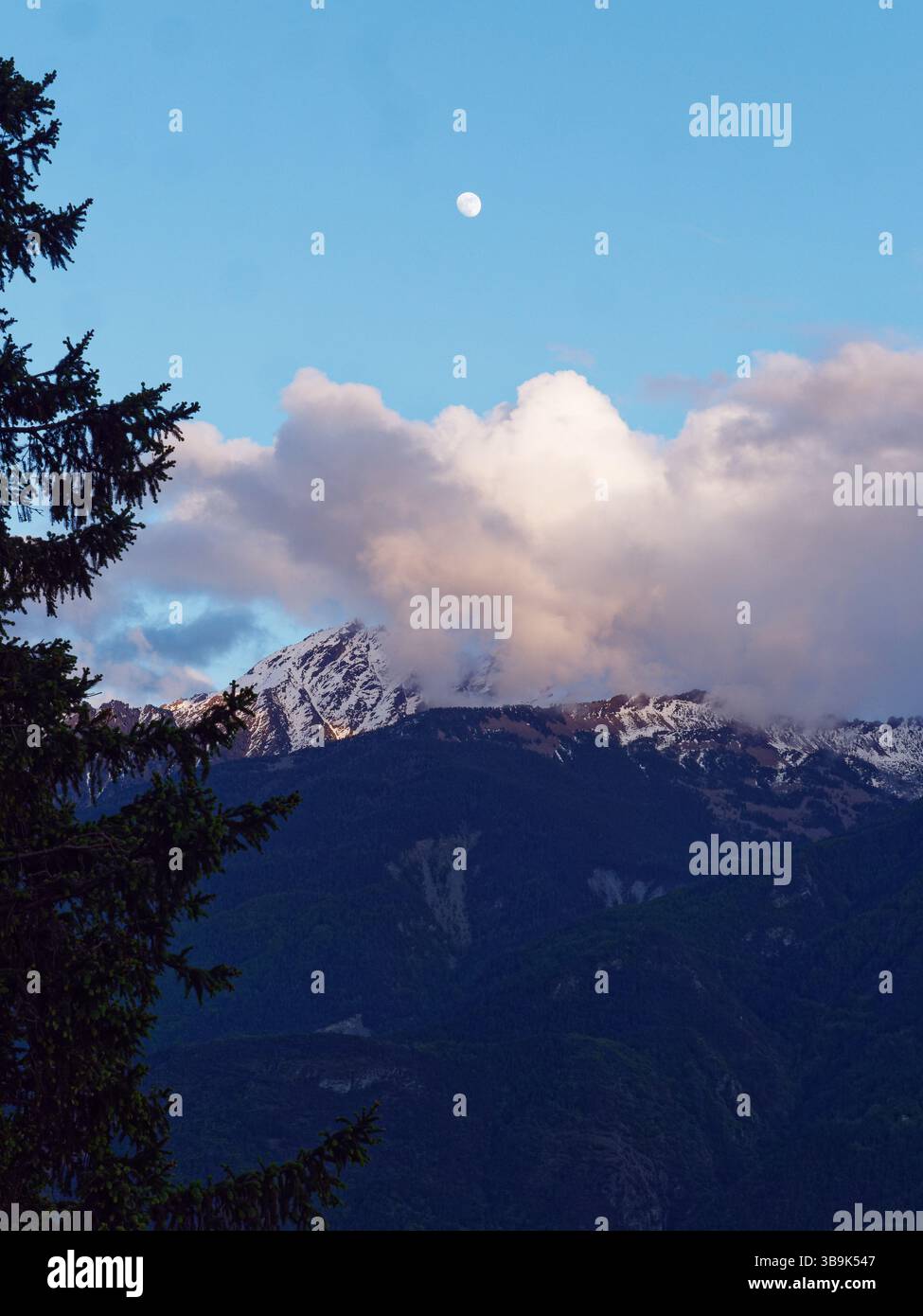Mond hoch in einem blauen Himmel über weißen Wolken, mit schneebedeckten Bergen darunter. Aosta Valley, NW Italien. Mai 2025 Stockfoto