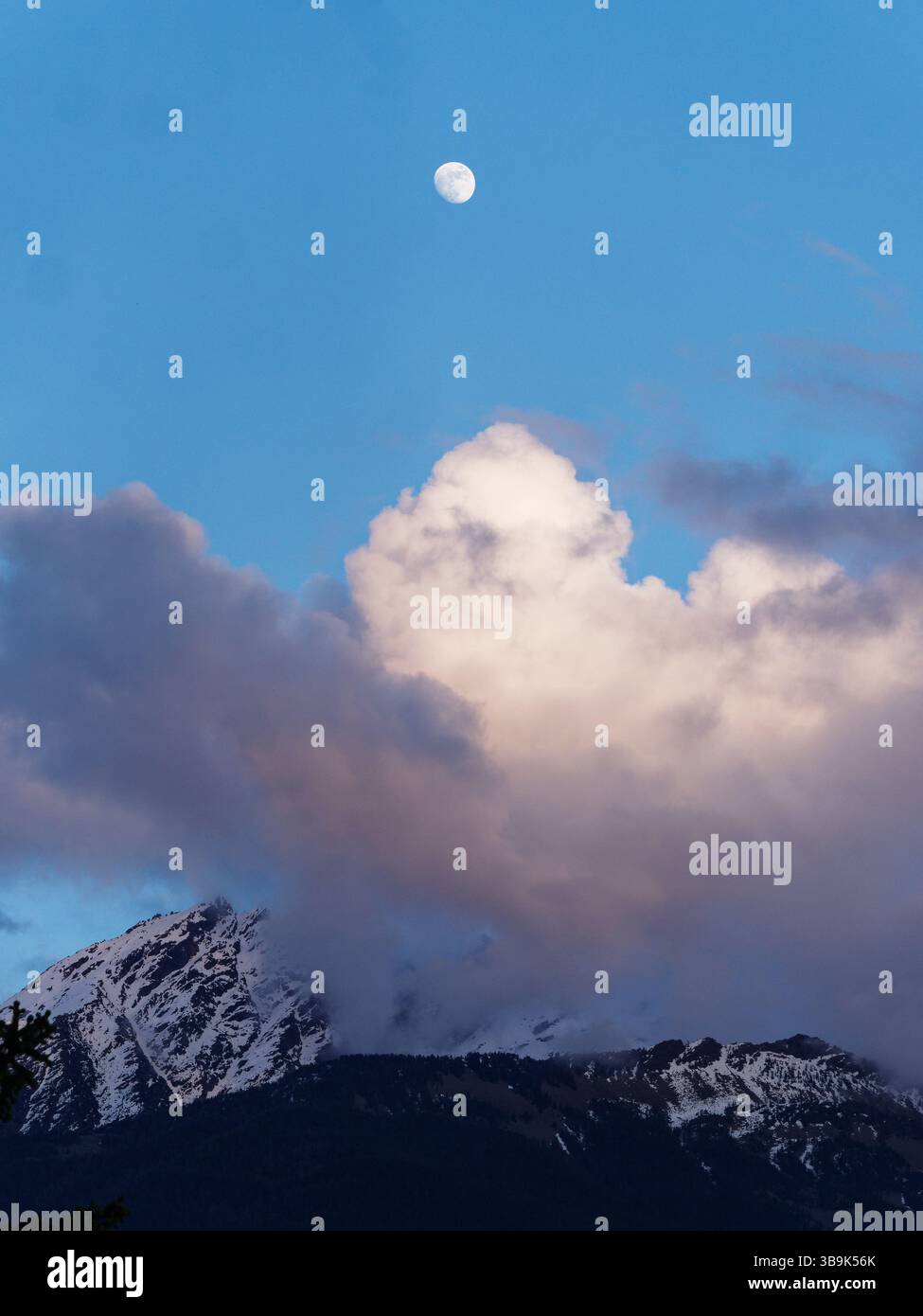 Mond hoch in einem blauen Himmel über weißen Wolken, mit schneebedeckten Bergen darunter. Aosta Valley, NW Italien. Mai 2025 Stockfoto