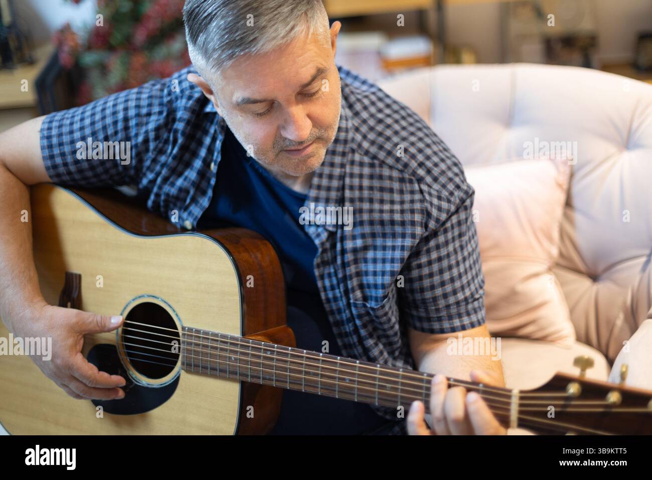 Porträt eines Reifen Mannes, der zu Hause in einem gemütlichen Stuhl sitzt und sich auf das Spielen einer Akustikgitarre konzentriert. Ein friedlicher Moment voller Hobbymusik Stockfoto