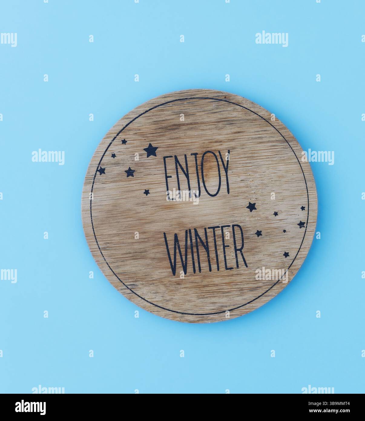 Ein runder Holzuntersetzer mit einem klaren und minimalistischen Design, in dem die Worte „Enjoy Winter“ in fetten Großbuchstaben eingeprägt sind. Stockfoto