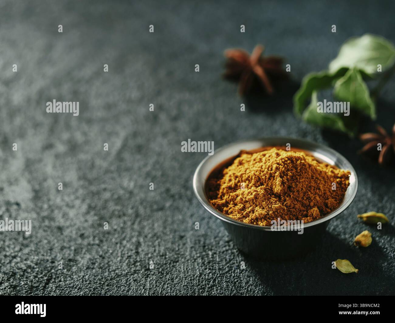 Indisches oder pakistanisches Masala-Pulver und Gewürze auf dunklem Strukturhintergrund. Kleine Metallschüssel mit trockenem, garam masala Mix Gewürzmischung, grüner Lorbeer Stockfoto