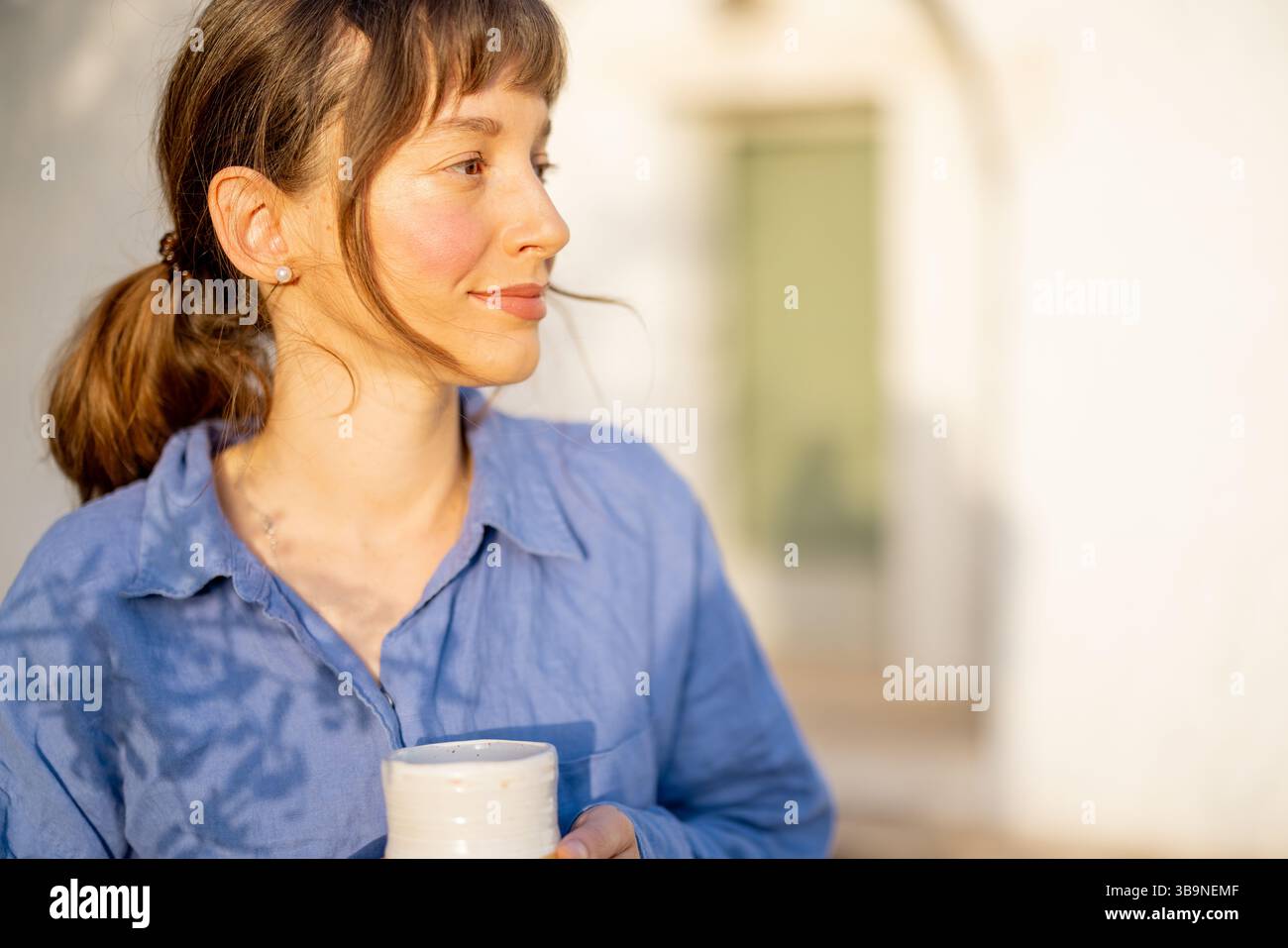 Nahaufnahme einer ruhigen Frau in blauem Leinen, die eine Keramikschale hält, in warmes Morgenlicht getaucht neben einem traditionellen trullo-Haus in Süditalien Stockfoto