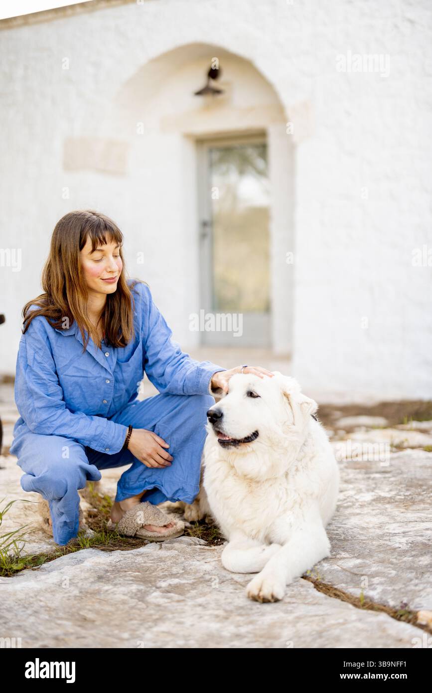 Eine Frau in blauem Outfit streichelt sanft ihren weißen Hund vor einem traditionellen trullo in Italien. Das Foto drückt Vertrauen, friedliche ländliche Lebensweise und emotionale Verbindung mit Tieren aus Stockfoto