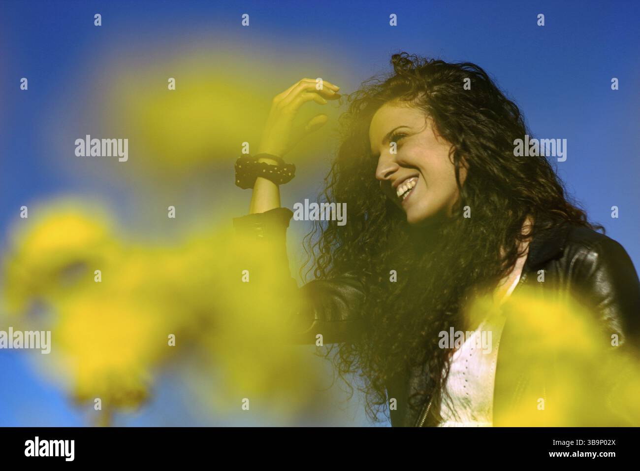 Porträt einer jungen Frau, die im Hintergrund lächelt, mit dem Vordergrund unscharfer gelber Blumen. Frühling Sommer Stockfoto