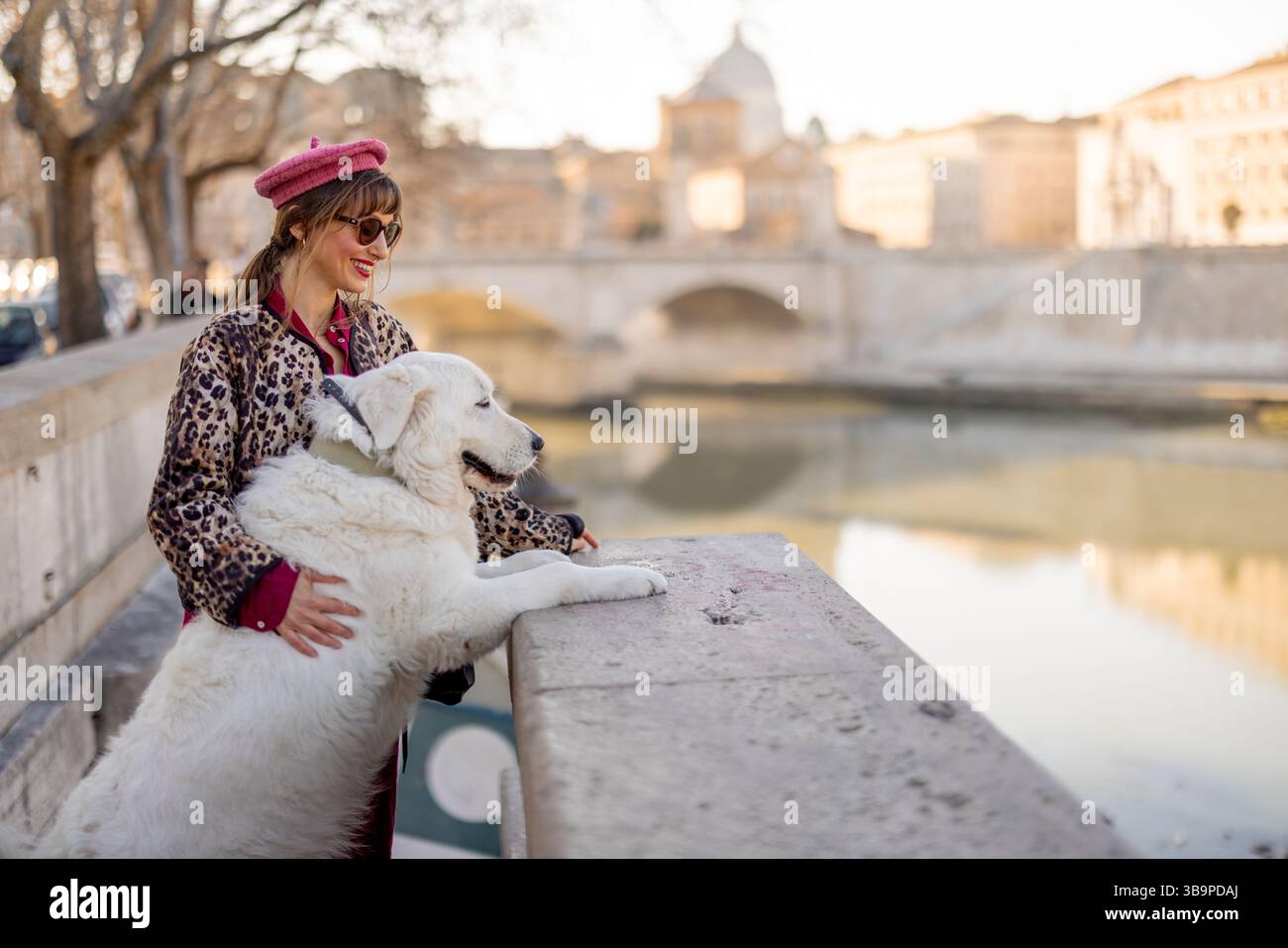 Eine Frau und ihr großer weißer Hund teilen einen zärtlichen Moment mit Blick auf den Fluss in Rom. Dieses Foto vermittelt Wärme und Mitgefühl und ist ideal für den Lebensstil von Haustieren und Reiseinhalte Stockfoto