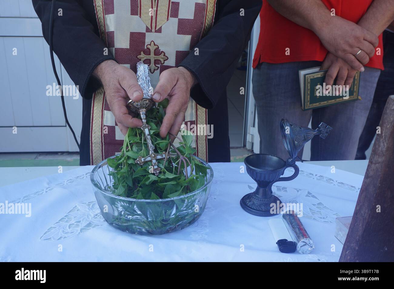 Ein Priester hält Basilikum und ein silbernes Kreuz über einer mit Weihwasser gefüllten Glasschale für die Einweihungszeremonie am ersten Schultag. Stockfoto