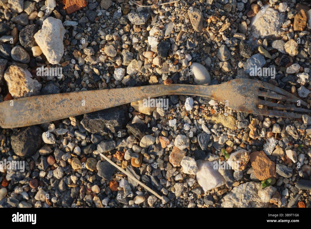 Eine verrostete Metallgabel liegt teilweise zwischen Kieseln und kleinen Felsen auf einer strukturierten Strandoberfläche vergraben. Stockfoto