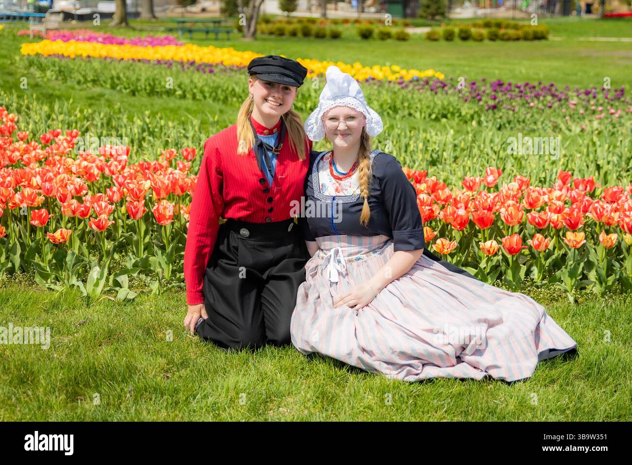 Holland, MI, USA - 24.04.2025: Porträt lächelnder Teenager-Mädchen in traditioneller niederländischer Kleidung, die an sonnigem Tag im blühenden Tulpenfeld stehen und Kultu zeigen Stockfoto