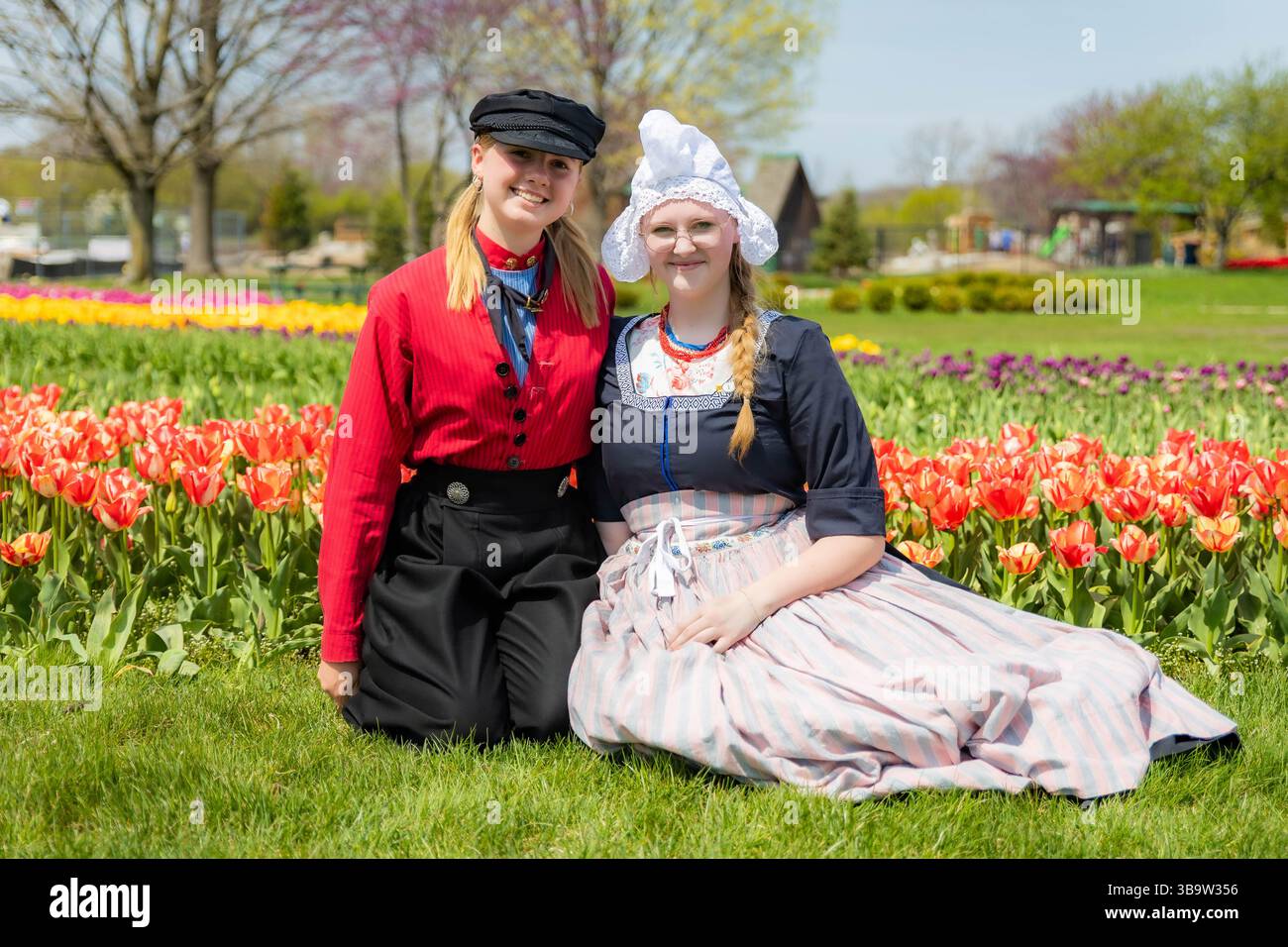 Holland, MI, USA - 24.04.2025: Porträt lächelnder Teenager-Mädchen in traditioneller niederländischer Kleidung, die an sonnigem Tag im blühenden Tulpenfeld stehen und Kultu zeigen Stockfoto