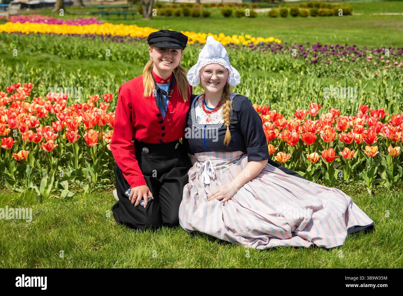 Holland, MI, USA - 24.04.2025: Porträt lächelnder Teenager-Mädchen in traditioneller niederländischer Kleidung, die an sonnigem Tag im blühenden Tulpenfeld stehen und Kultu zeigen Stockfoto