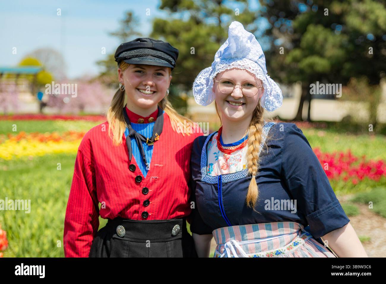 Holland, MI, USA - 24.04.2025: Porträt lächelnder Teenager-Mädchen in traditioneller niederländischer Kleidung, die an sonnigem Tag im blühenden Tulpenfeld stehen und Kultu zeigen Stockfoto