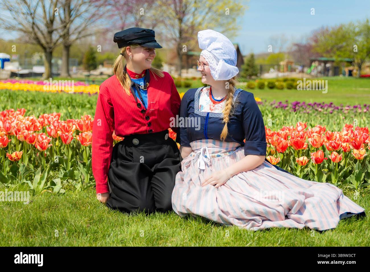 Holland, MI, USA - 24.04.2025: Porträt lächelnder Teenager-Mädchen in traditioneller niederländischer Kleidung, die an sonnigem Tag im blühenden Tulpenfeld stehen und Kultu zeigen Stockfoto