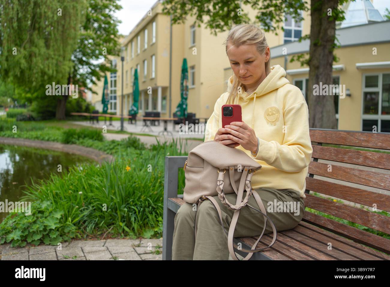 Frau in einem gelben Kapuzenpullover, die auf einer Parkbank sitzt und auf einem Smartphone SMS schreibt. Sie wirkt konzentriert und engagiert, mit einem entspannten Ausdruck, umgeben von Stockfoto