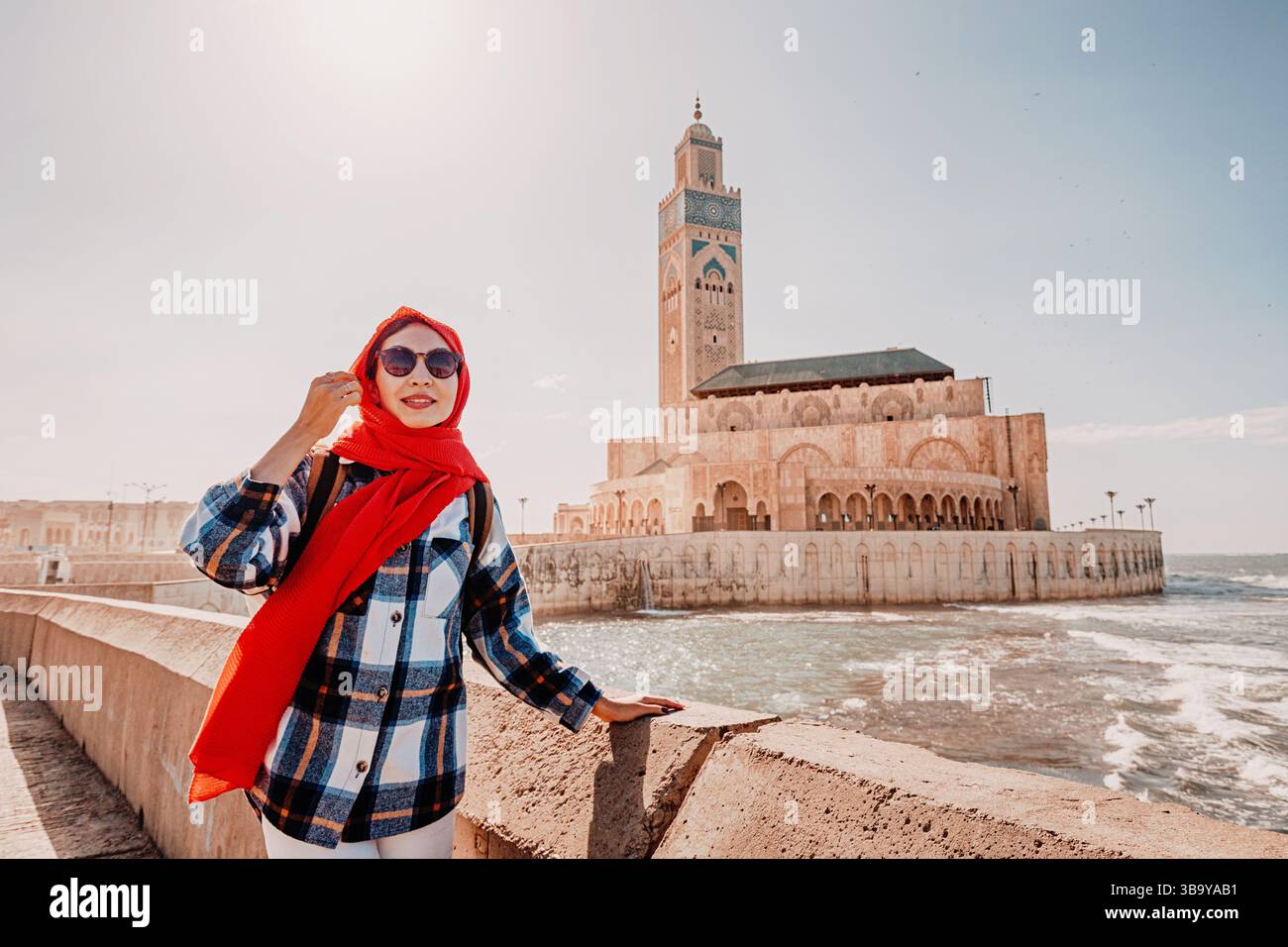 Junge Frau mit rotem Schal und Sonnenbrille posiert vor der Moschee Hassan II. In Casablanca, Marokko Stockfoto