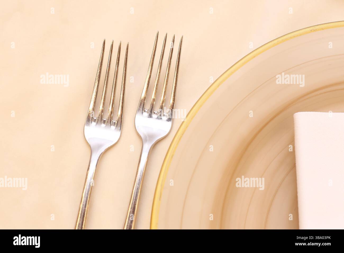 Elegante Tischdekoration. Zwei silberne Gabeln auf beigefarbenem Hintergrund. Glasplatte mit goldenem Rand. Sanfte Pastelltöne. Gute Küche. Stockfoto