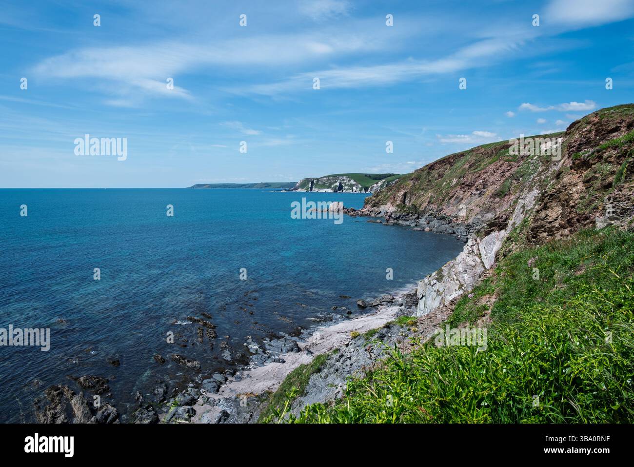 Ein malerischer Blick auf die Küste, mit den zerklüfteten Klippen, die unter einem hellen, teilweise bewölkten Himmel auf das ruhige blaue Meer treffen. Stockfoto