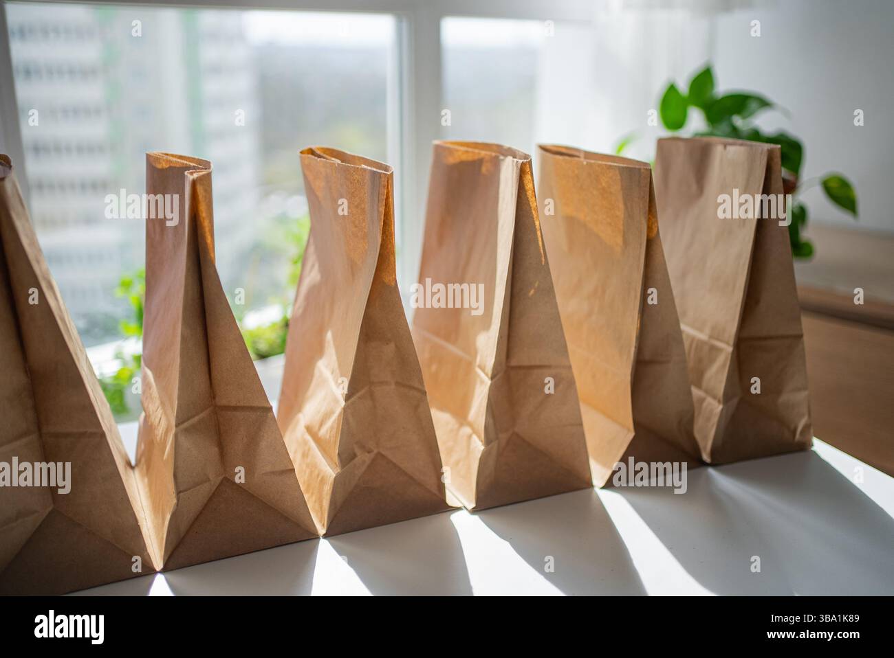 Mehrere braune Papiertüten stehen offen auf einem weißen Tisch, die mit Lebensmitteln für die Lieferung gefüllt werden können, beleuchtet durch Sonnenlicht von einem nahe gelegenen Fenster Stockfoto
