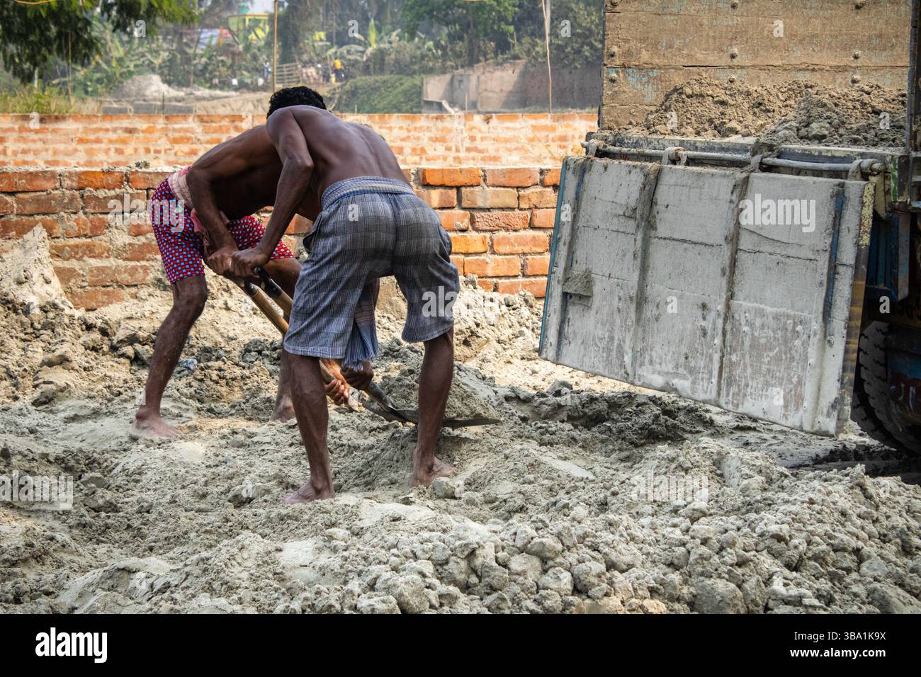 Zwei Männer, die mit Schaufeln Sand von einer Baustelle in der Nähe eines Müllwagens transportieren Stockfoto