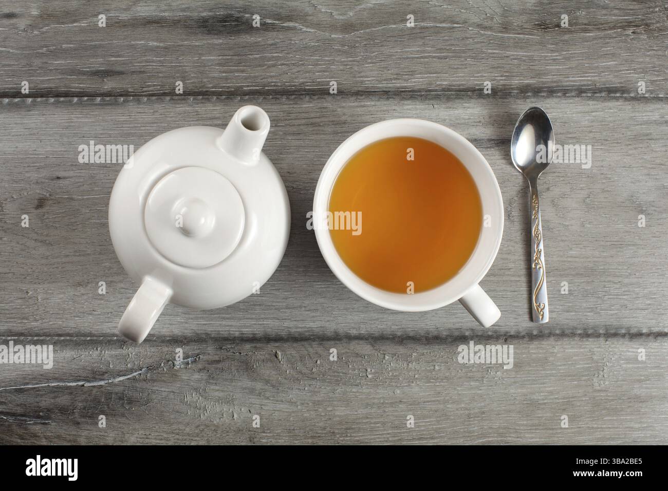 Blick von der Tischplatte auf weiße Keramik-Teekanne, eine Tasse heißem schwarzen Tee und einen silbernen Löffel auf einem grauen Holztisch Stockfoto