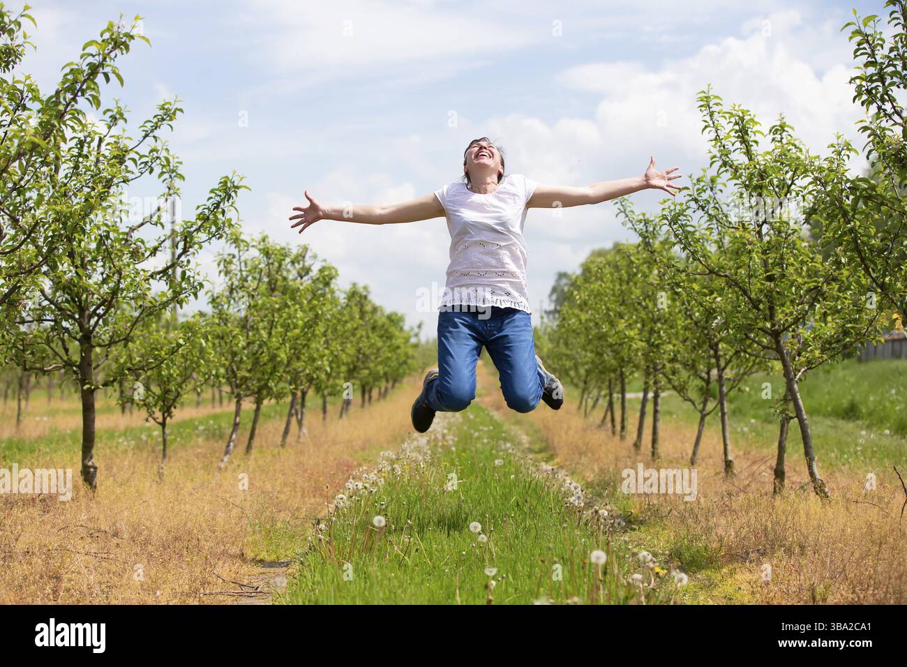 Eine erwachsene Frau jubelt und springt vor der Kulisse eines bluehenden Fruehlingsgartens auf Stockfoto
