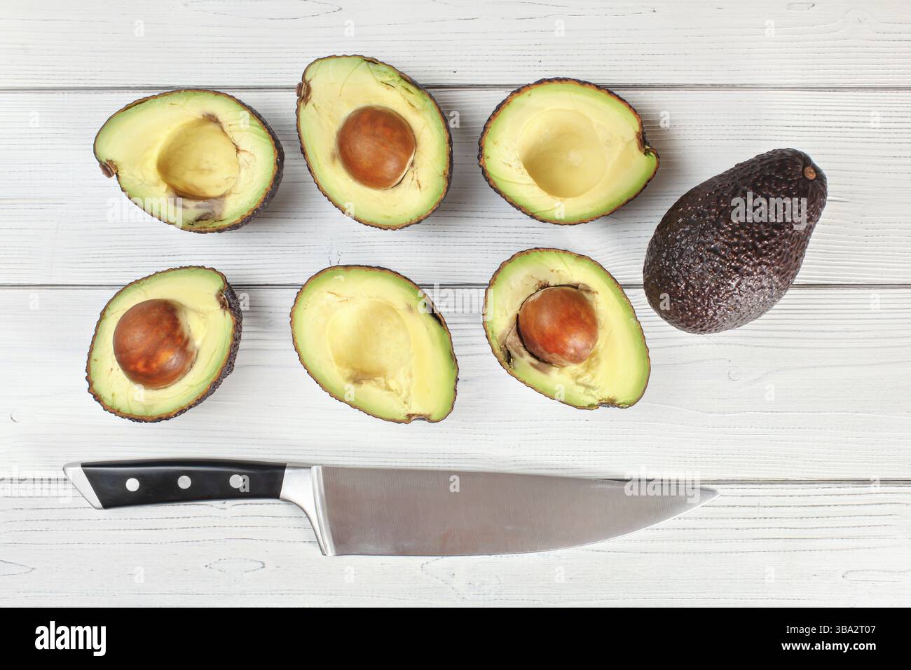Avocadohälften und ein ganzes Obst auf weißem Bretttisch, darunter ein Kochmesser. Blick von oben, Liptovsky Hradok, Slowakei, Europa Stockfoto