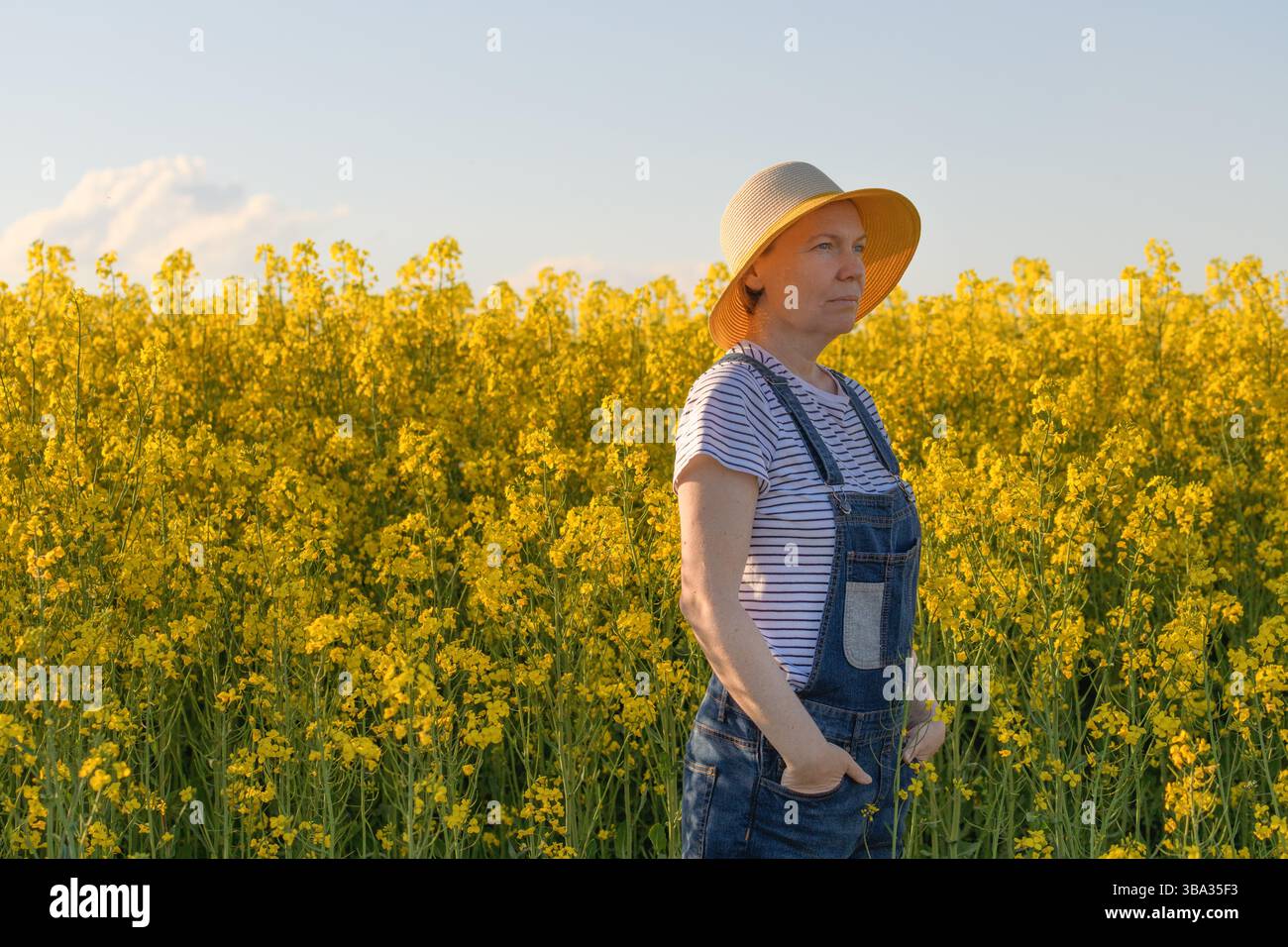 Porträt einer schönen erwachsenen Farmerin mit Strohhut, die am sonnigen Frühlingnachmittag auf einem blühenden Rapsplantafeld steht, selektiver Fokus Stockfoto